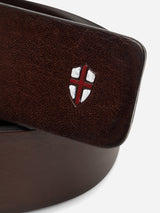 A close-up of the BGathol Belt - Dark Brown by Blue de Gênes, handmade in Italy from full-grain leather, showcases a classic metal buckle and a belt loop adorned with a small shield emblem featuring a red cross on a white background.