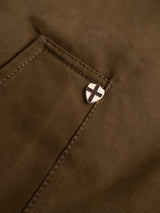 A close-up of the BGjacques AW Coat in Brown by Blue de Gênes shows the heavy cotton fabric pocket with precise stitching, accented near the top edge by a metallic shield-shaped pin adorned with a black cross—a signature workwear-inspired detail.