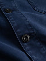 Close-up of the BGcuddy Nebraska Jacket - Blue by Blue de Gênes, featuring a navy blue fabric with black buttons, distinct visible stitching, and neat buttonholes. The smooth, slightly reflective material is reminiscent of the high-density dobby weave characteristic of classic utility jackets.