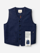 The BGlumen Nebraska Gilet - Blue by Blue de Gênes is a navy blue button-up workwear waistcoat with two front pockets, shown flat against a white background. Made from high-density cotton featuring a subtle dobby weave, this vest offers both durability and style. A product tag, attached to the right pocket, displays text alongside a small illustration of the gilet.