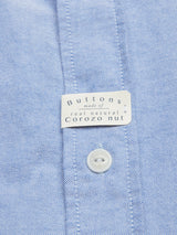 A close-up of the BGsandro Oxford Shirt - Blue by Blue de Gênes, made from light blue organic cotton, features a white label stating "Buttons made of real natural Corozo nut" and one visible white button below the label.