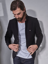 A man with long hair and a beard wears the Casella Officers Blazer in black from Blue de Gênes over a light gray shirt and dark jeans, looking down as he buttons his jacket while standing against a plain light-colored wall.