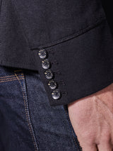 A close-up of a person’s wrist shows the sleeve of the Blue de Gênes Casella Officers Blazer in black, detailed with five black buttons, alongside part of a pair of blue denim jeans.