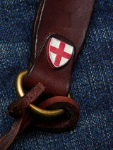 A close-up of the Blue de Gênes BGsavino Key Hanger - Brown, crafted from cow leather and featuring a brass ring and a red cross emblem on a white shield, resting on blue denim fabric.