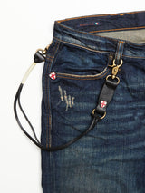 A pair of blue denim jeans with distressed details, featuring a Blue de Gênes BGsavino Key Hanger in black—with metal clasps attached to the belt loop—and small red-and-white cross patches near the pocket.