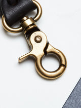 A close-up of the BGsavino Key Hanger - Black by Blue de Gênes, featuring a brass swivel clasp attached to a black leather strap, resting on a white and gray background.