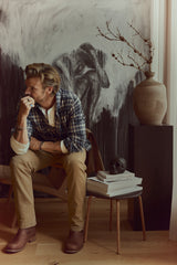 A man wearing the BGenrico Hang Shirt - Navy Check by Blue de Gênes and khaki pants sits on a chair, resting his chin on his hand. Nearby are a vase with dried branches, a black skull, and a stack of books in front of a dark abstract painting.
