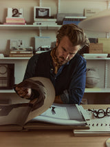 A man with light brown hair and a scarf, dressed in the Blue de Gênes BGgregor Denim Shirt in Denim Blue, looks through a large book at a desk, surrounded by books and objects on softly lit shelves. Glasses and another open book rest on the table nearby.