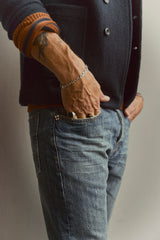 A person wearing a navy jacket, Blue de Gênes’ BGdaw Stripe Knit in Ginger Bread Orange, and blue jeans stands with one hand in their pocket, displaying a tattoo, bracelet, and ring.