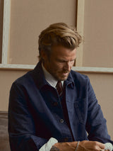 A man with light brown hair and a beard looks down, wearing a navy jacket over a white shirt and the BGcarl Tie - Ginger Bread Orange by Blue de Gênes, seated indoors against a beige background with framed panels.
