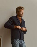 A man with tousled hair stands against a plain light wall, wearing the Blue de Gênes BGlewis Wool Blazer in Dark Navy over a brown and orange striped sweater and blue jeans, looking thoughtfully to the side.
