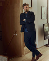 A man with a beard and mustache stands indoors with arms crossed, dressed in a dark suit paired with the BGpalco Ian Pant - Black by Blue de Gênes, featuring a relaxed fit and tapered silhouette. He wears a blue shirt and brown boots, posing near a staircase and TV in a modern, warmly lit room.