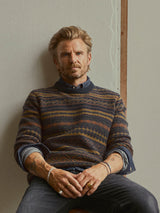 A man with blond hair and a beard sits against a plain wall, wearing the Blue de Gênes BGrhys Knit sweater in Dark Navy over a collared shirt, his sleeves rolled up and jewelry adorning his wrists and fingers, looking directly at the camera.