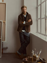 A man with blond hair and a beard stands by a large window in a studio filled with art supplies and canvases. He wears a brown jacket, white shirt, and BGleroy Jeans - Raw Denim from Blue de Gênes, paired with boots, his arms crossed as he surveys the space.