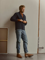 A man with light brown hair and a beard stands against a beige wall, wearing a navy jacket, striped sweater, and Blue de Gênes BGgiacomo Alfie Jeans in mid blue denim—relaxed loose fit jeans made of comfort stretch denim—paired with brown boots. A framed item leans nearby on the floor.