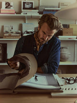 A man with light brown hair and a beard, dressed in a three-piece suit featuring the BGduncan Ian Gilet in Black from Blue de Gênes, sits at a desk flipping through a large book, surrounded by shelves filled with books and objects in a softly lit room.