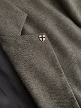 A close-up of the BGFukui Wool Blazer - Grey Mel by Blue de Gênes showcases an unstructured pocket adorned with a shield-shaped pin featuring a cross design. The fabric’s subtle diagonal pattern in earthy grey melange tones highlights the effortless sophistication and relaxed fit of this wool blazer.