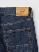 Close-up of the back pocket on BGmarco Cast Jeans - Denim wash by Blue de Gênes, crafted from cotton denim in blue with yellow stitching and finished with a tan leather patch above the pocket featuring a stamped shield emblem.