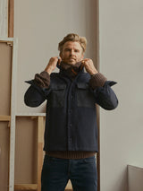 A man with light hair and a beard stands indoors, adjusting the collar of a Blue de Gênes BGBoyd Wool Jacket in Dark Navy over a brown sweater. He looks slightly up and forward, with unfinished canvas frames leaning against a wall behind him.