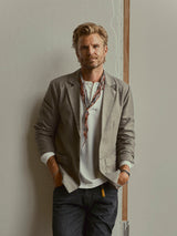A man with blonde hair and a beard stands against a wall, dressed in the Blue de Gênes BGFukui Wool Blazer in Grey Mel over a white shirt and scarf, paired with dark jeans. His hands appear to be amputated at the wrists.