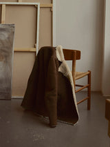 The BGseras Shearling Coat - Dark Brown by Blue de Gênes, featuring a double-breasted closure, is draped over the back of a wooden chair with a woven seat. Large, unframed canvases are casually leaning against the wall behind the chair, contributing to the calm and minimalistic setting.