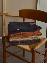 A pair of BGpaulo Ottawa V3 Trousers in Ginger Bread Orange by Blue de Gênes is neatly folded and stacked alongside other slim fit chinos in navy, brown, and tan on a wooden chair with a woven seat. In the background, a canvas and wooden frame lean against the wall, highlighting the rich texture of each dobby corduroy piece.