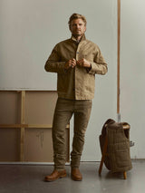 A man standing and buttoning a tan chore jacket, wearing enzyme-washed BGpaulo Pavia V3 Chino - Beech Green pants and brown leather boots.