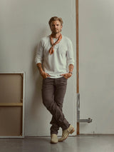 A man wearing a white henley, an orange patterned neckerchief, and brown Blue de Gênes BGvinci Iro Pant - Hot Fudge Brown leans against a wall.