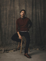 A man wearing a ginger bread orange Blue de Gênes BGhamish Crewneck Knit, dark trousers, and a neckerchief sits on a wooden stool.