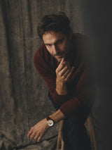 A man wearing a Blue de Gênes BGhamish Crewneck Knit in Ginger Bread Orange sits on a wooden stool, resting his chin on his hand.