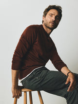 A man wearing a brown long-sleeve sweater over a white shirt and charcoal Blue de Gênes BGpaulo Pavia 1 V2 Chino trousers sits on a wooden stool.