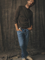 A man wearing a long-sleeved brown sweater, Blue de Gênes BGvinci Pala Used V3 Jeans - Denim Blue with a brown belt, and light-colored sneakers.
