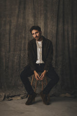 A man wearing a dark jacket, white t-shirt, and Blue de Gênes BGvinci Black OD V3 Jeans - Blue Black Denim sits on a wooden stool.