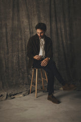 A man wearing a dark jacket and white shirt sits on a wooden stool, wearing Blue de Gênes BGvinci Black OD V3 Jeans in blue black denim.