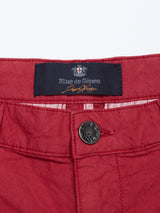 Close-up of Teo Milano Shorts - Havana Red by Blue de Gênes, showcasing an embossed button and dark waistband label with white text and a small crossed elements logo. Made from a soft, lightweight cotton/linen blend.