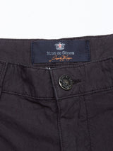 Close-up of Teo Milano Shorts in Jet Black by Blue de Gênes, featuring a navy label with brand text and a stitched signature. The waistband is fastened with a metallic button engraved with Blue de Gênes. The cotton/linen blend fabric has a textured look with visible stitching around the pockets.