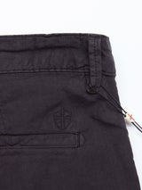 Close-up of Jet Black Teo Milano Shorts by Blue de Gênes, made from a soft cotton/linen blend with an embroidered shield emblem, side pocket, and a tag with a red cross near the waistband, emphasizing its smooth texture and visible stitching.