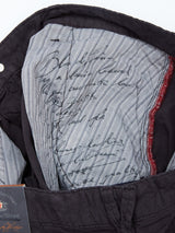 The image showcases the interior lining of the Teo Milano Shorts by Blue de Gênes, featuring a blue striped fabric, suggesting a refined cotton/linen blend. The fabric has handwritten black text, and the exterior is jet black with subtle red stitching on a seam.