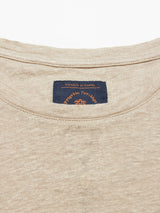 Close-up of the Totti T-Shirts neckline in pebble beige linen, showing a dark blue Blue de Gênes label with orange Progetto Heritage text and emblem. The fabric has a natural texture, with Le realtà di Genova at the top of the label.