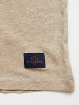 Close-up of the Totti T-Shirt by Blue de Gênes in Pebble Beige, featuring a navy rectangular patch with an embroidered red-orange shield logo and Blue Bones text. Made from 100% Linen Jersey, the shirts natural texture and neatly stitched edges create a clean hemline.