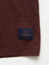 Close-up of the Totti T-Shirt - Prune by Blue de Gênes, featuring a brown, natural linen texture with a small dark blue rectangular patch embroidered with a shield design and light orange text near the bottom edge.