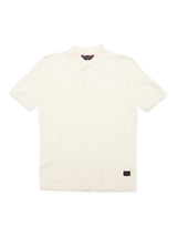 The Primo Polo Knit - Ecru by Blue de Gênes is a cream-colored polo with a cotton/linen blend. It has short sleeves, a classic collar, and a three-button placket, plus a small black logo patch near the bottom hem for added flair.