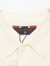 A close-up of the Primo Polo Knit - Ecru shows a cream-colored cotton/linen blend collar with a button detail. The inner label bears the Blue de Gênes name and small shield logo, featuring a blue and brown color scheme.