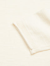 A close-up of the cream-colored Monte Knit - Whitecap by Blue de Gênes shows its neatly folded fabric on a matching background. The texture, resembling a cotton/linen blend, reveals slight ridges and patterns, featuring an elegant sewn roll-rib hem on the edge.