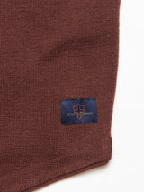 Close-up of a brown knit Zorba waistcoat fabric with a dark label featuring an orange cross design and the text true genes. Made from Merino wool, the texture is noticeable, with the Blue de Gênes label stitched on the lower right corner.