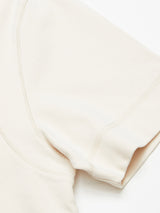 Close-up of the sleeve and shoulder seam of a collarless Nipote AW S/S Grandad - Whitecap t-shirt by Blue de Gênes, in light beige on a white surface. The soft cotton interlock jersey fabric has a slightly textured appearance with visible stitching along the edges.