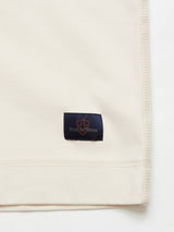A close-up of the cream-colored Nipote AW S/S Grandad - Whitecap by Blue de Gênes shows cotton interlock jersey fabric with a small black patch at the hem. The patch has a subtle logo and blue sense in orange, embodying the classic style of a Grandad T-Shirt with neatly stitched edges.