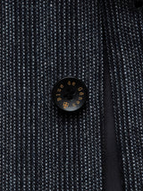 Close-up of a textured dark gray fabric with a black button, displaying Blue de Gênes in gold letters. This woven pattern, crafted from an Italian cotton-wool blend, showcases exquisite Portuguese tailoring reminiscent of the Canna Dori Gilet - Grey stripe.