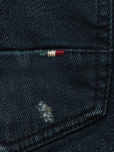 A close-up of Vinci Haworth Jeans - Denim Blue by Blue de Gênes shows Comfort Stretch Denim with a torn patch and subtle red, white, and green stitches. The texture highlights the masterful handmade retouching, showcasing a small area of wear and repair.