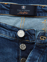 Close-up of Blue de Gênes Repi Scan Used Jeans in mid blue denim, showing the label stitched inside the waistband. Noticeable details are a metallic button embossed with the brands name, intricate pocket stitching, all made from comfort stretch denim for ease and style.